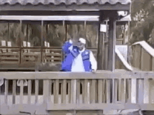 a man in a blue jacket is standing on a balcony with his hand up .