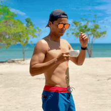 a shirtless man wearing sunglasses and a baseball cap stands on a sandy beach