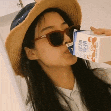 a woman wearing sunglasses and a straw hat drinks from a carton of milk