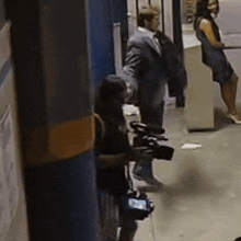 a man in a suit is holding a camera in a hallway while a woman stands behind him