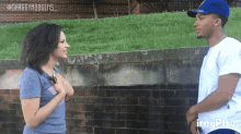 a man and a woman are standing in front of a brick wall and the man is wearing a dodgers hat