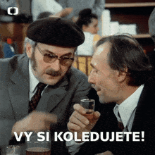 two men sitting at a table with vy si koledujete written in the corner