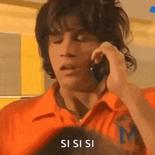 a man in an orange shirt is talking on a cell phone and making a funny face .