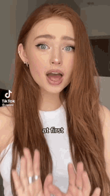a woman with long red hair and blue eyes is wearing a white tank top and making a funny face .