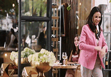 a woman in a pink jacket is dancing in front of a restaurant while another woman sits at a table .