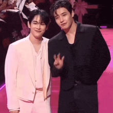 two men are standing next to each other on a pink carpet and one is giving a peace sign .