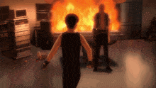 a man in a black tank top stands in front of a fire with a man holding a gun behind him
