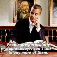 a man in a suit and tie is sitting at a desk reading a book and talking on a cell phone