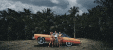 a group of people standing in front of an orange car with palm trees in the background