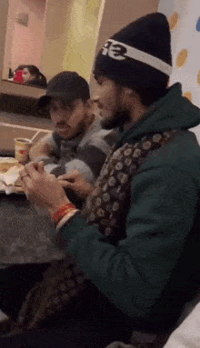 two men are sitting at a table with one wearing a beanie that says te on it
