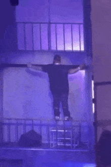 a man is standing on a balcony with his arms outstretched and a purple light behind him .