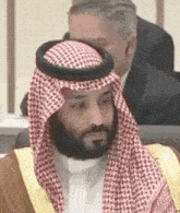 a man with a beard wearing a keffiyeh and a robe is sitting in a chair .