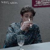a man sitting at a table drinking from a wine glass with a wildcards logo in the background