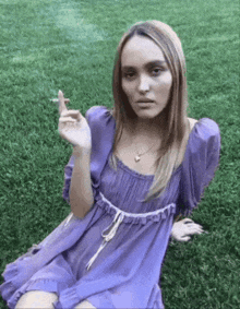 a woman in a purple dress is smoking a cigarette in the grass .