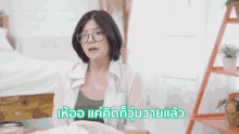 a woman wearing glasses is sitting in front of a bed with the words " แค่ คิด ก็ รู้ ว่า " written in green