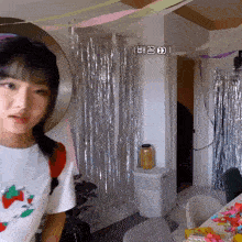 a girl in a white shirt with strawberries on it is standing in a room with silver tinsel on the wall
