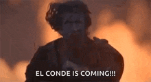 a man is standing in front of a fire with the words `` el conde is coming '' .