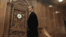 a man in a tuxedo stands in front of a clock that shows the time as almost 5:00