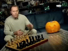 a man sits on a couch playing a game of chess next to a carved pumpkin that says tht on it