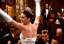 a woman in a white dress and tiara is raising her arms in the air in a crowded room .