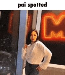 a woman in a white sweater is standing in front of a neon sign that says pai spotted