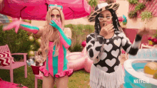 two women are standing next to each other in front of a pool and a pink umbrella .