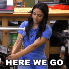 a woman in a blue jumpsuit is standing in front of a wooden table with the words here we go above her