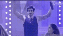 a man in a black tank top is standing in front of a purple wall with his arms in the air .