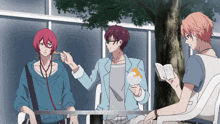 a man with red hair is feeding another man a spoon while another man reads a book
