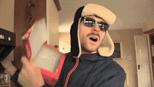 a man wearing a furry hat and sunglasses is holding a red box