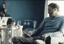 a man is sitting in a chair next to a baby in a crib while using a tablet