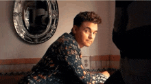 a young man in a floral shirt is sitting in front of a mirror in a room .
