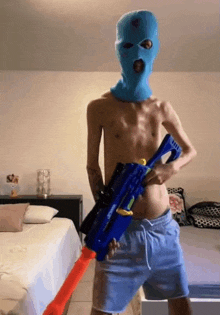 a shirtless man wearing a blue ski mask is holding a toy gun in a bedroom .