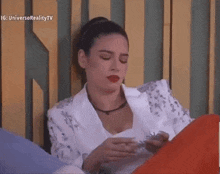 a woman in a white jacket and red lipstick is sitting on a bed holding something in her hands .