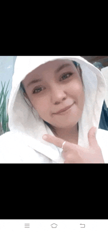 a woman wearing a white hoodie is smiling and giving a peace sign .