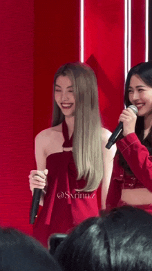 a woman in a red dress holds a microphone while another woman smiles in front of a red wall