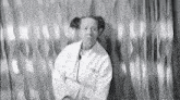 a black and white photo of a woman in a white coat standing in front of a wall with her arms crossed .