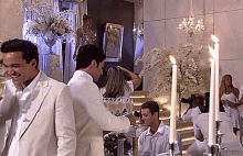 a man in a white suit stands in a room with candles and a woman in a silver dress
