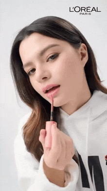 a woman wearing a white hoodie is applying lipstick by l'oreal paris