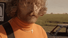 a man with a beard and glasses wears an orange turtleneck and a gold cross necklace