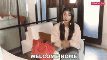 a woman is sitting on a couch with a red purse and the words welcome home behind her