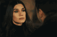 a man with a beard wearing a black turtleneck and a black coat is talking to another man .