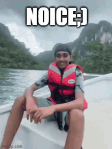 a young man wearing a life jacket is sitting on a boat with mountains in the background and the caption noice