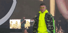 a man wearing a neon yellow hoodie and sunglasses stands in front of a large screen