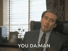 a man in a suit and tie is smoking a cigarette and saying `` you da man '' while sitting in a chair .