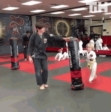 a man in a black karate uniform is teaching a young boy how to kick a century punching bag .
