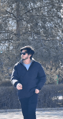 a man wearing sunglasses and a black jacket is walking in a park