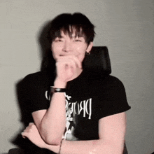 a man wearing a black t-shirt is covering his mouth with his hands while sitting in a chair .
