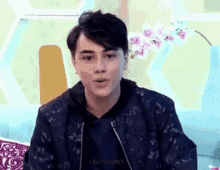 a young man in a black jacket is sitting on a couch talking to someone .