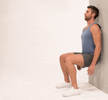 a man is squatting against a wall wearing a blue tank top and grey shorts